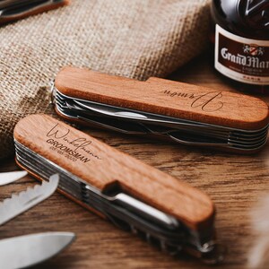 Peut inclure: Deux couteaux de poche &agrave; manche en bois avec du texte grav&eacute;. Un couteau porte le texte "Wickelmann GROOMSMAN 08.06.2020" et l'autre porte le texte "monur A".