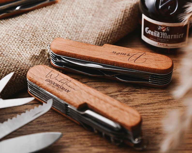 Peut inclure: Deux couteaux de poche &agrave; manche en bois avec du texte grav&eacute;. Un couteau porte le texte "Wickelmann GROOMSMAN 08.06.2020" et l'autre porte le texte "monur A".