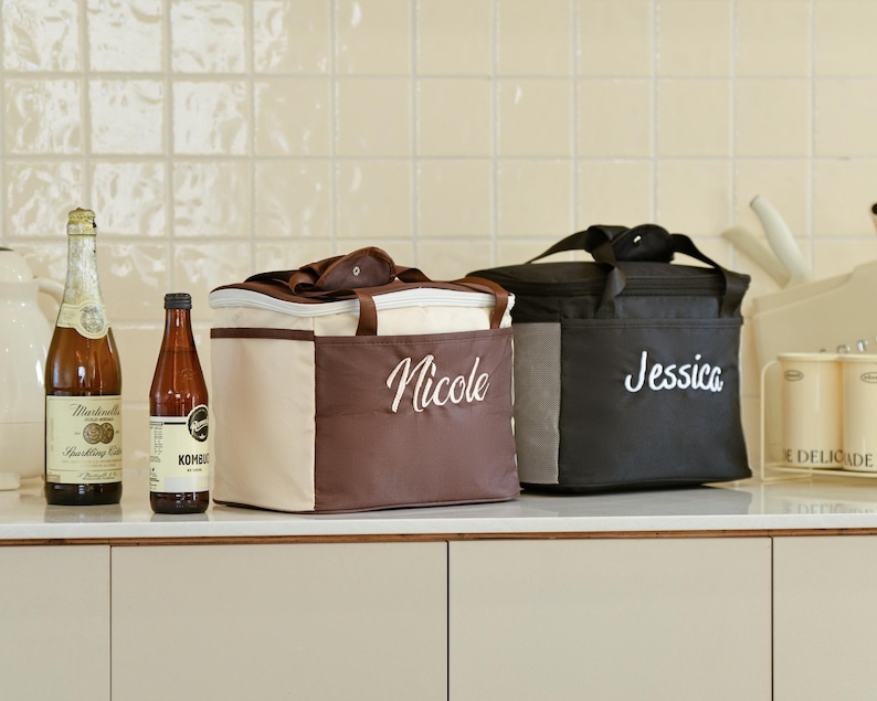 Pu&ograve; includere: Due borse da pranzo isolate in tessuto marrone e nero. La borsa a sinistra ha il nome "Nicole" ricamato. La borsa a destra ha il nome "Jessica" ricamato.