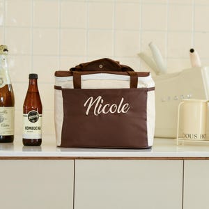 Pu&ograve; includere: Una borsa termica per il pranzo marrone e beige con chiusura a zip. La borsa ha un'etichetta bianca con il nome "Nicole" ricamato. In primo piano ci sono due bottiglie di vetro, una con un'etichetta che dice "Kombucha".