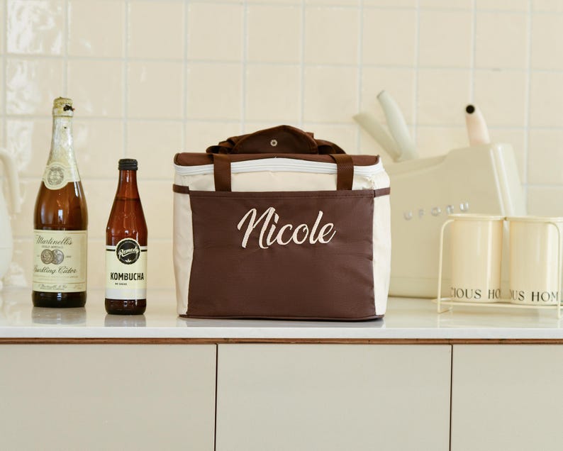 Pu&ograve; includere: Una borsa termica per il pranzo marrone e beige con chiusura a zip. La borsa ha un'etichetta bianca con il nome "Nicole" ricamato. In primo piano ci sono due bottiglie di vetro, una con un'etichetta che dice "Kombucha".