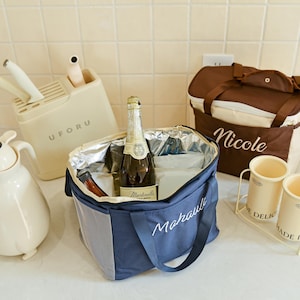 Pu&ograve; includere: Una borsa termica blu e bianca con la parola "Marauli" ricamata. La borsa &egrave; piena di ghiaccio e una bottiglia di vino spumante. C'&egrave; una borsa termica marrone e beige con la parola "Nicole" ricamata. Ci sono due contenitori in ceramica bianca con le parole "Delic" e "Made in" su di essi.