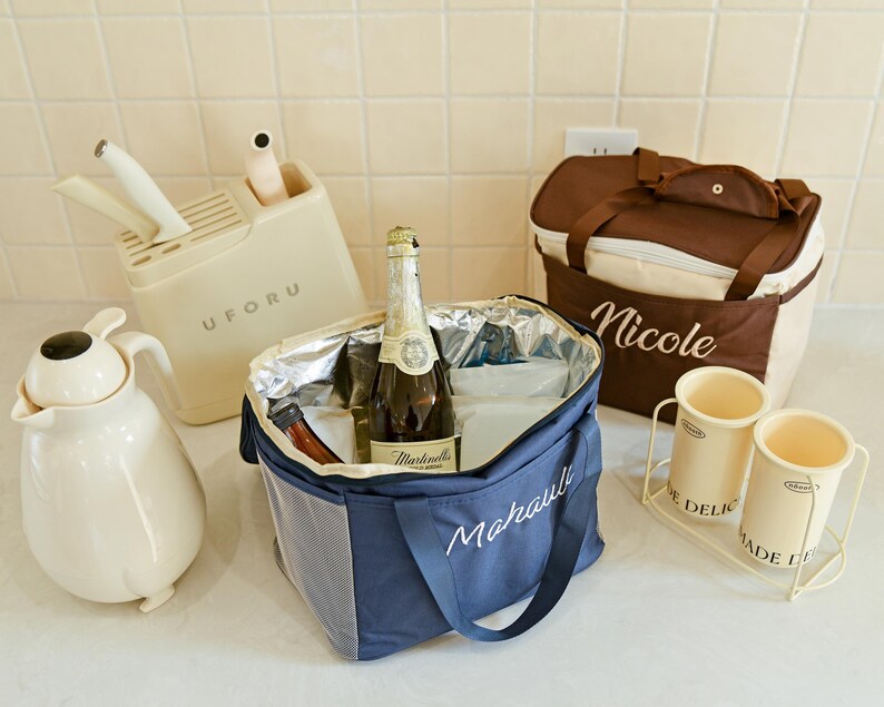 Pu&ograve; includere: Una borsa termica blu e bianca con la parola "Marauli" ricamata. La borsa &egrave; piena di ghiaccio e una bottiglia di vino spumante. C'&egrave; una borsa termica marrone e beige con la parola "Nicole" ricamata. Ci sono due contenitori in ceramica bianca con le parole "Delic" e "Made in" su di essi.