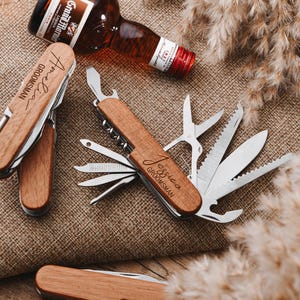 Peut inclure: Deux couteaux de poche multi-outils &agrave; manche en bois avec les mots "Groomman" et "Jessica Groomman" grav&eacute;s sur les manches. Les couteaux ont plusieurs outils, dont un ouvre-bouteille, une scie, un tournevis et une lame de couteau.