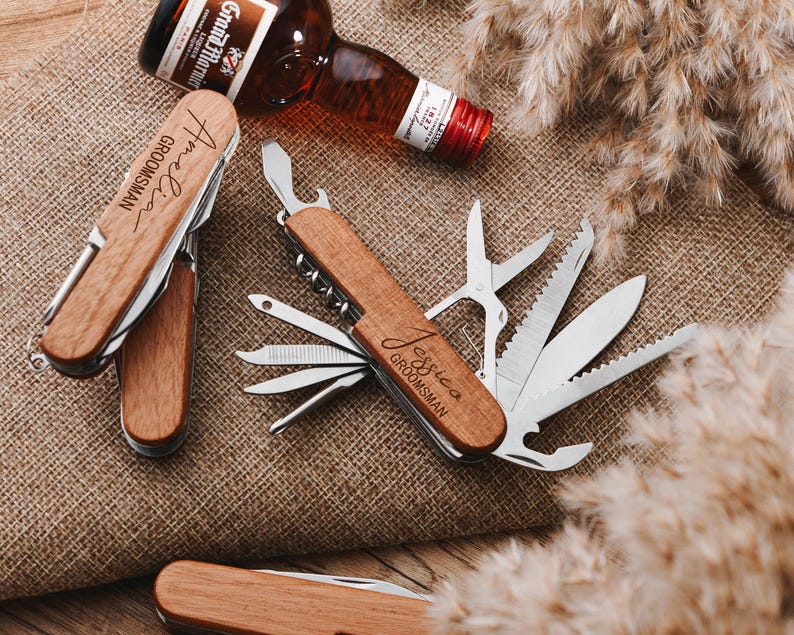 Peut inclure: Deux couteaux de poche multi-outils &agrave; manche en bois avec les mots "Groomman" et "Jessica Groomman" grav&eacute;s sur les manches. Les couteaux ont plusieurs outils, dont un ouvre-bouteille, une scie, un tournevis et une lame de couteau.