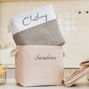 Peut inclure: Deux paniers de rangement en tissu avec des poignées en corde. Le panier du haut est gris avec des lettres blanches qui disent "Clothing". Le panier du bas est beige avec des lettres blanches qui disent "Sundries".