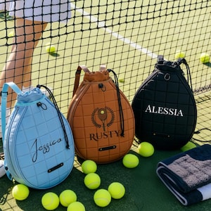 May include: Three tennis ball-shaped bags in blue, brown, and black, each with a name on the front. The blue bag says "Jessica", the brown bag says "Rusty", and the black bag says "Alessia". Tennis balls and a towel are also visible.