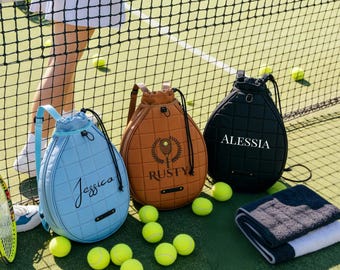 May include: Three tennis ball-shaped bags in blue, brown, and black, each with a name on the front. The blue bag says "Jessica", the brown bag says "Rusty", and the black bag says "Alessia". Tennis balls and a towel are also visible.