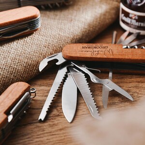 Peut inclure: Un couteau de poche en bois avec une inscription personnalis&eacute;e qui dit "[Name]  Groom'sman 09.09.2020". Le couteau poss&egrave;de une vari&eacute;t&eacute; d'outils, dont un ouvre-bouteille, une scie, un tournevis et une paire de ciseaux.