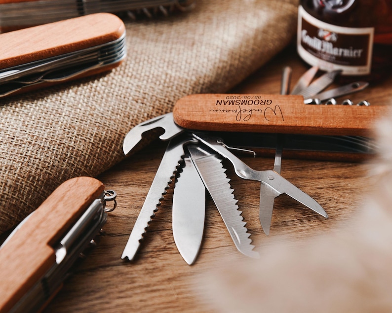 Peut inclure: Un couteau de poche en bois avec une inscription personnalis&eacute;e qui dit "[Name]  Groom'sman 09.09.2020". Le couteau poss&egrave;de une vari&eacute;t&eacute; d'outils, dont un ouvre-bouteille, une scie, un tournevis et une paire de ciseaux.