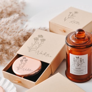 May include: Two gift boxes with personalized floral designs and the names "Katie" and "Natalie" on them. A brown glass jar with a lid and a label that says "Lavender Scented Candle" is in the foreground.
