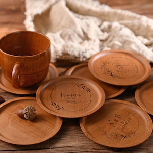 May include: Set of eight wooden coasters with personalized engravings. Each coaster features a floral design and a unique name and date. The coasters are a light brown color and have a smooth, polished finish.