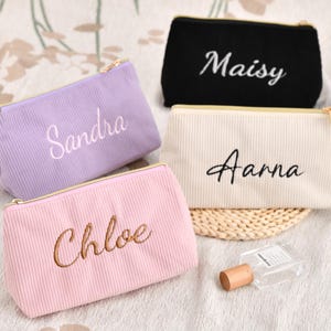 Può includere: Quattro trousse per cosmetici in velluto a coste dai colori pastello: lavanda, rosa, crema e nero. Ogni trousse ha una cerniera dorata e un nome ricamato in un colore complementare: Sandra, Chloe, Aarna e Maisy. È visibile anche un piccolo flacone di profumo.