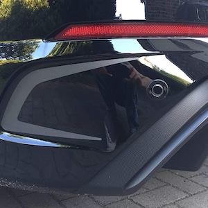 May include: Black car rear bumper with a black trim piece and a rear parking sensor. The bumper has a textured finish.