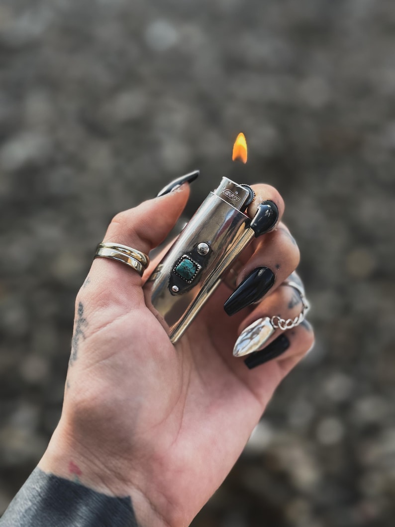 May include: A silver lighter with a turquoise stone inlay and a flame coming out of the top. The lighter is being held by a hand with black nail polish.