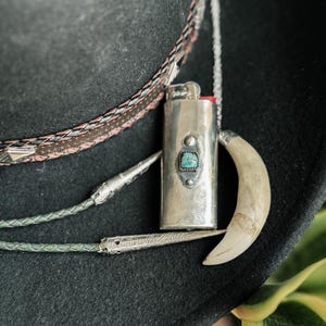May include: A silver lighter with a turquoise stone inlay hangs from a chain on a black cowboy hat. The lighter is attached to a silver and leather bolo tie.