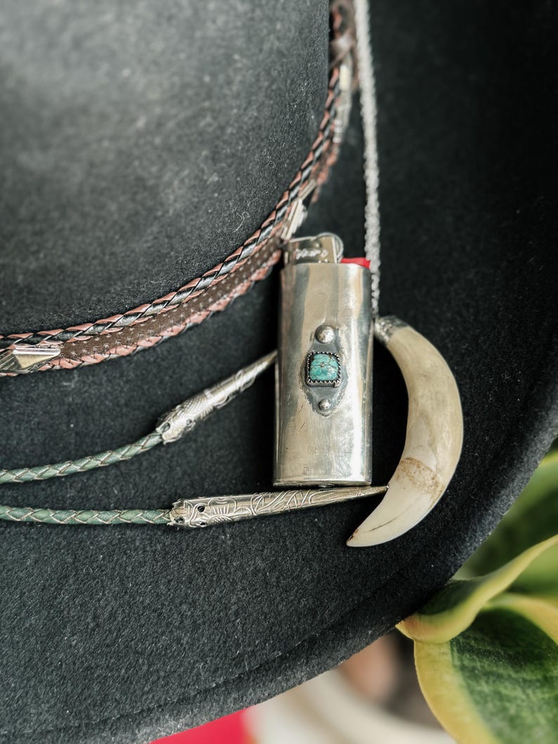 May include: A silver lighter with a turquoise stone inlay hangs from a chain on a black cowboy hat. The lighter is attached to a silver and leather bolo tie.