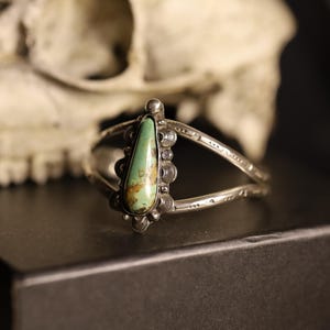 May include: A silver cuff bracelet with a teardrop-shaped turquoise stone. The stone is light green with brown inclusions. The bracelet has a split band design and decorative silver accents. The background includes a skull.