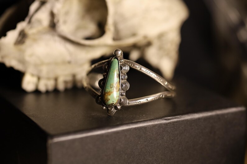 May include: A silver cuff bracelet with a teardrop-shaped turquoise stone. The stone is light green with brown inclusions. The bracelet has a split band design and decorative silver accents. The background includes a skull.