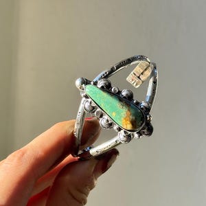 May include: A silver-toned ring featuring a large, teardrop-shaped turquoise stone. The ring has intricate metalwork around the stone and a decorative band. The turquoise stone has a mix of green and brown hues.