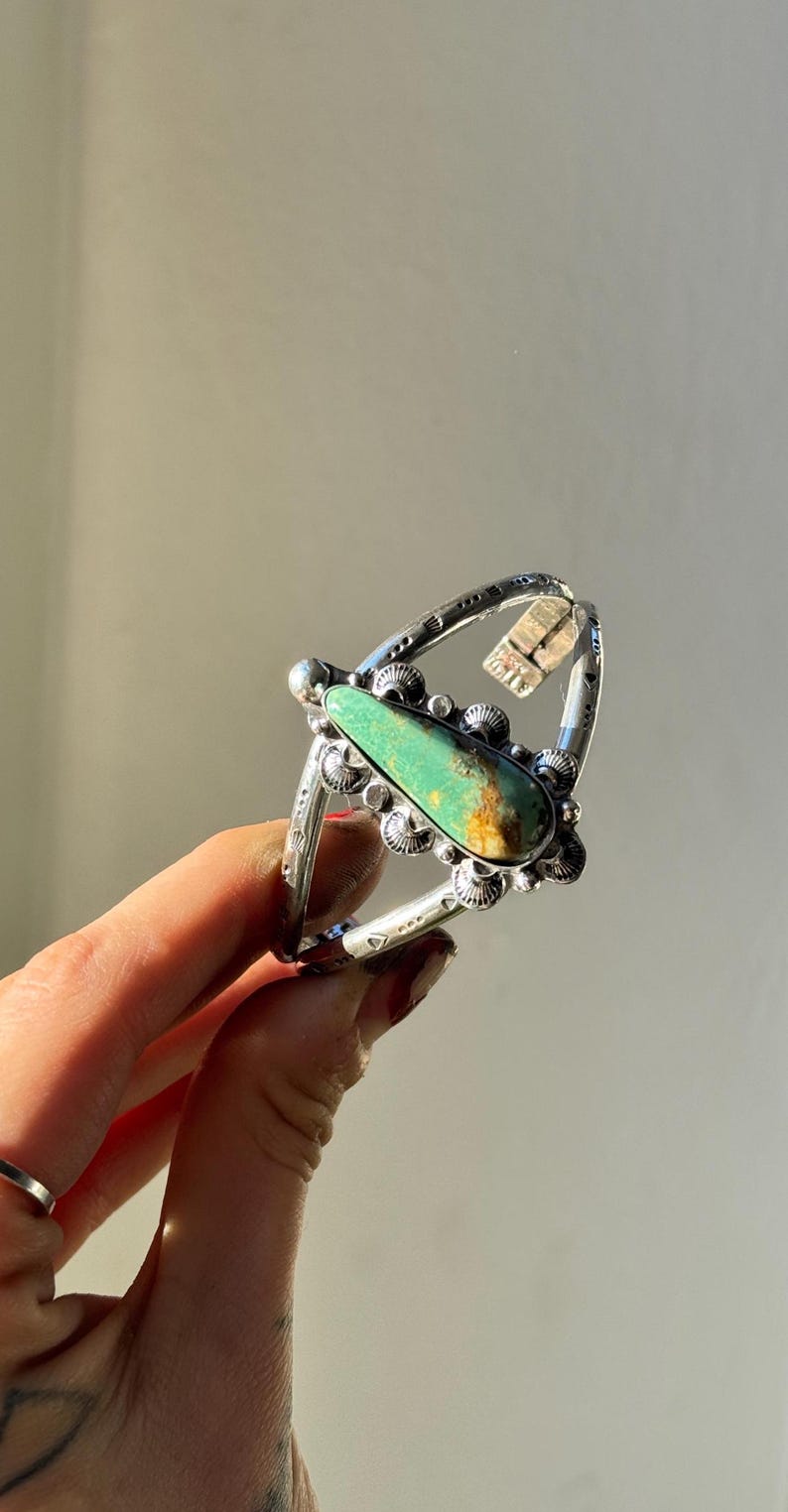 May include: A silver-toned ring featuring a large, teardrop-shaped turquoise stone. The ring has intricate metalwork around the stone and a decorative band. The turquoise stone has a mix of green and brown hues.