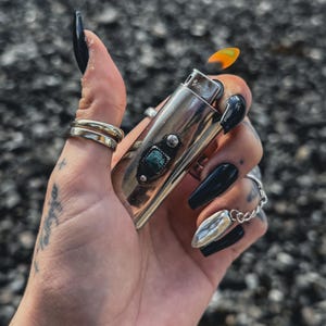 May include: A silver lighter with a turquoise stone inlay and a flame coming out of the top. The lighter is being held by a hand with black nail polish.