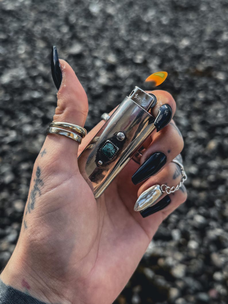 May include: A silver lighter with a turquoise stone inlay and a flame coming out of the top. The lighter is being held by a hand with black nail polish.