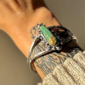 May include: A silver cuff bracelet with a large, oval turquoise stone. The stone has brown and green hues. The bracelet is worn on a tattooed arm, with a beige knit sleeve visible.