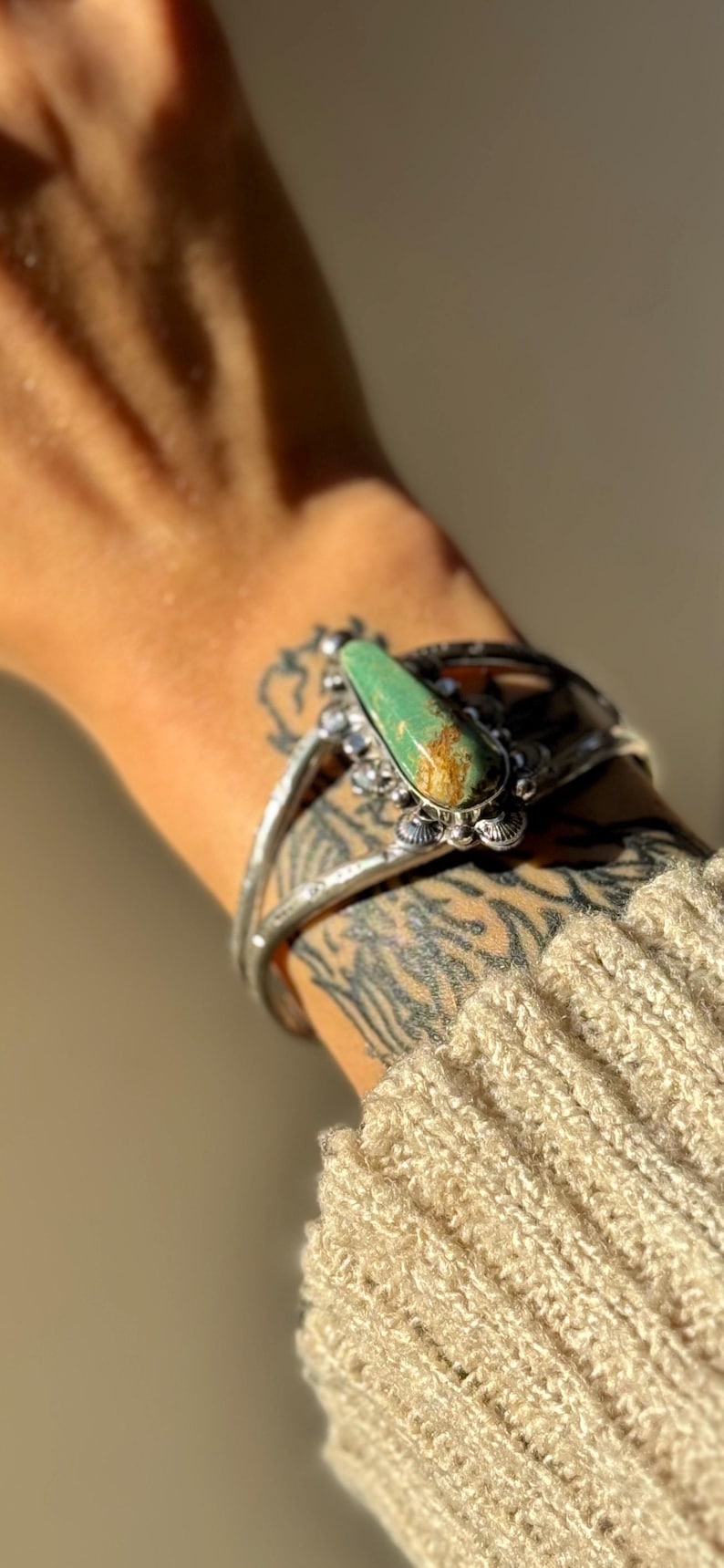 May include: A silver cuff bracelet with a large, oval turquoise stone. The stone has brown and green hues. The bracelet is worn on a tattooed arm, with a beige knit sleeve visible.