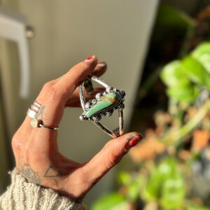 May include: A silver cuff bracelet with a large, teardrop-shaped turquoise stone at the center. The turquoise is a vibrant green with brown matrix. The bracelet has a detailed, ornate design with small silver beads and decorative elements.