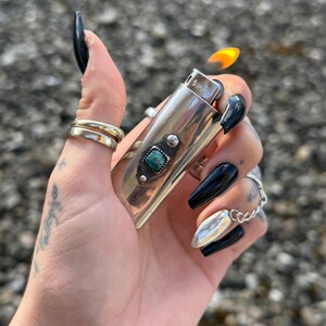 May include: Silver lighter with a turquoise stone inlay and a flame coming out of the top. The lighter is being held by a hand with black nail polish.
