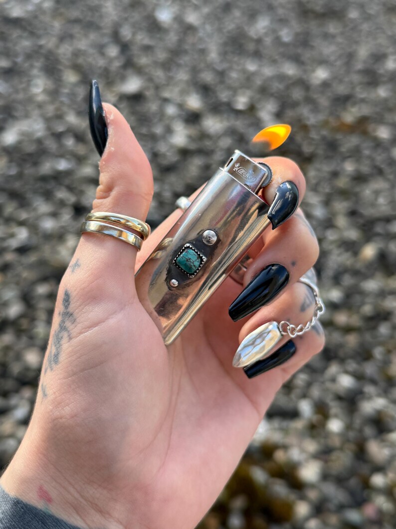May include: Silver lighter with a turquoise stone inlay and a flame coming out of the top. The lighter is being held by a hand with black nail polish.