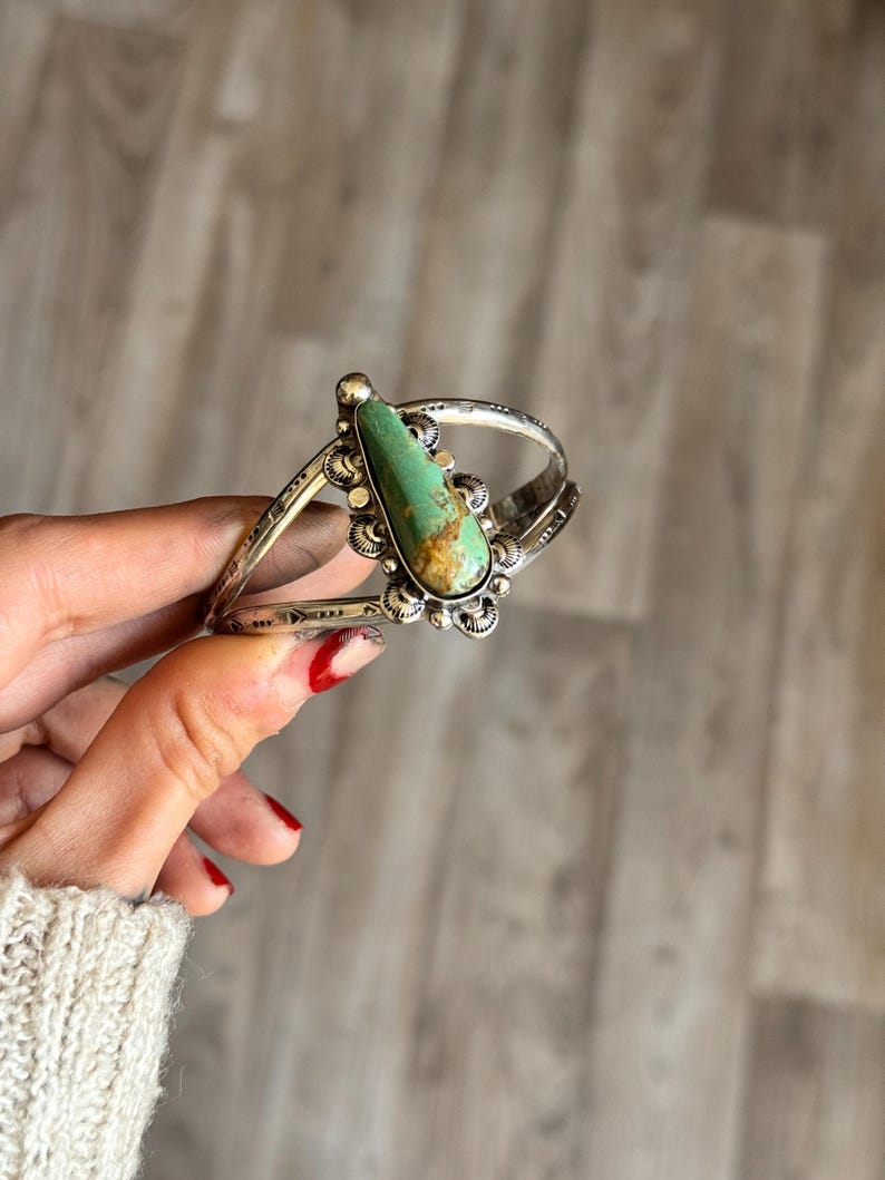 May include: A silver cuff bracelet featuring a large, teardrop-shaped turquoise stone. The stone is set in a decorative bezel with a floral design. The bracelet has etched details and a split band design.