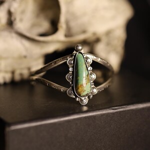 May include: Silver cuff bracelet with a teardrop-shaped turquoise stone. The stone is a vibrant green with brown marbling. The bracelet has an open design with decorative detailing and is set against a dark background.