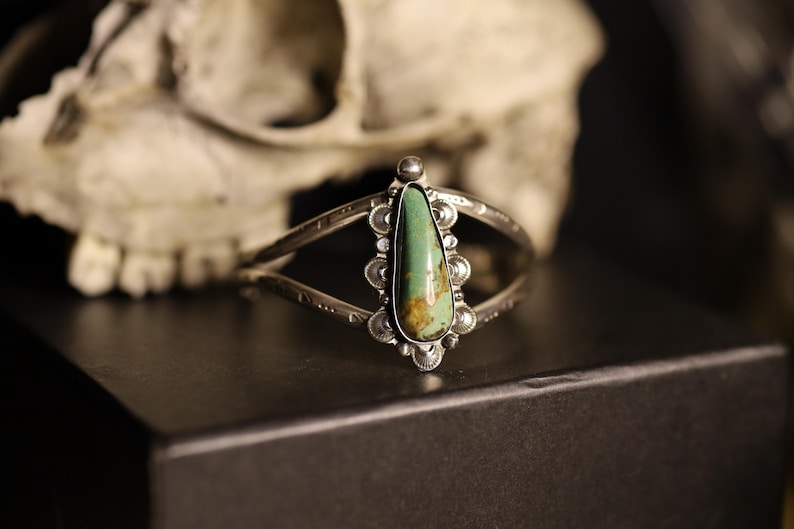 May include: Silver cuff bracelet with a teardrop-shaped turquoise stone. The stone is a vibrant green with brown marbling. The bracelet has an open design with decorative detailing and is set against a dark background.