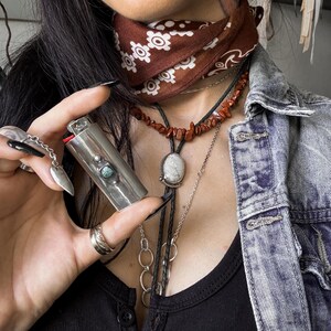 May include: A silver lighter with a turquoise stone inlay, held in a hand wearing a silver ring. The person is wearing a brown bandana with white floral pattern, a red coral necklace, and a silver necklace with a white stone pendant.