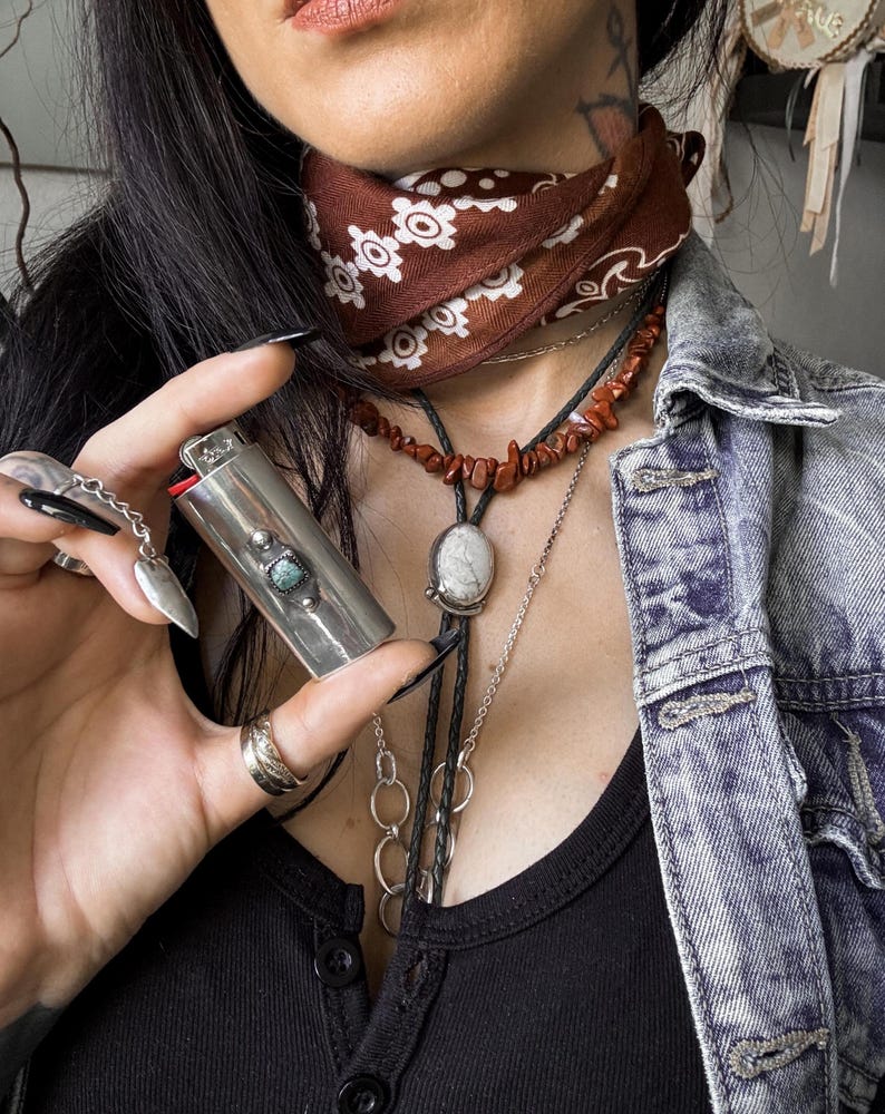 May include: A silver lighter with a turquoise stone inlay, held in a hand wearing a silver ring. The person is wearing a brown bandana with white floral pattern, a red coral necklace, and a silver necklace with a white stone pendant.
