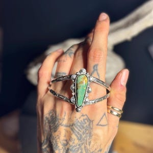 May include: Silver cuff bracelet featuring a large, teardrop-shaped turquoise stone. The bracelet has intricate stamped designs and decorative silver accents. A unique piece of jewelry.