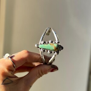 May include: Silver cuff bracelet featuring a large, teardrop-shaped turquoise stone. The bracelet has an open design with intricate metalwork and decorative elements. The turquoise stone is a vibrant green with brown matrix.