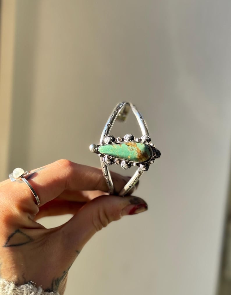 May include: Silver cuff bracelet featuring a large, teardrop-shaped turquoise stone. The bracelet has an open design with intricate metalwork and decorative elements. The turquoise stone is a vibrant green with brown matrix.