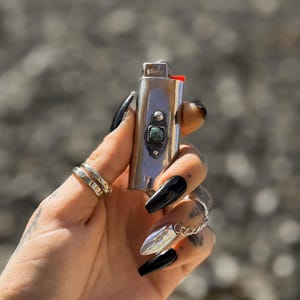 May include: A silver lighter with a turquoise stone inlay. The lighter is held in a hand with black nail polish.