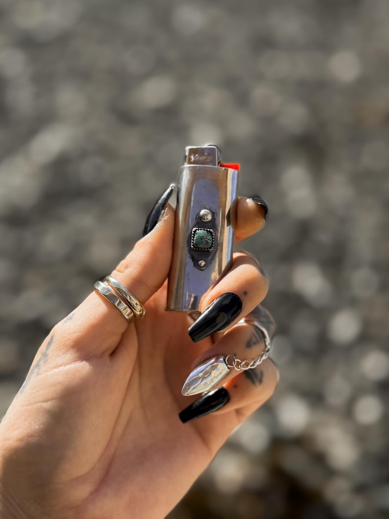 May include: A silver lighter with a turquoise stone inlay. The lighter is held in a hand with black nail polish.