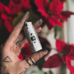May include: A silver lighter with a green stone inlay. The lighter is being held in a hand with a tattoo.