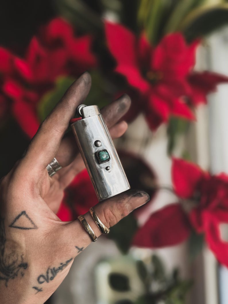 May include: A silver lighter with a green stone inlay. The lighter is being held in a hand with a tattoo.