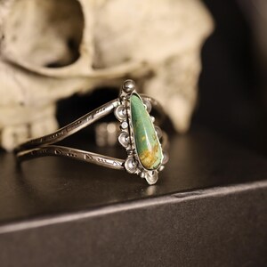 May include: Silver cuff bracelet with a teardrop-shaped turquoise stone. The bracelet has an open design with etched details and decorative accents around the stone. The background includes a skull.