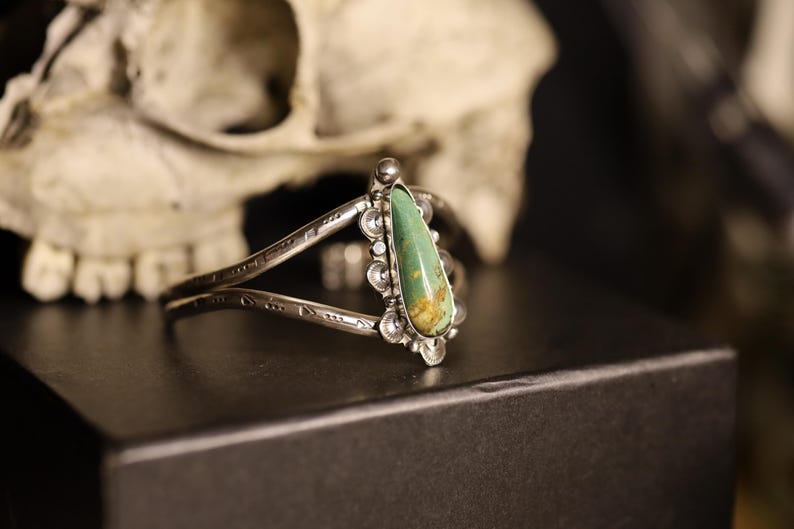 May include: Silver cuff bracelet with a teardrop-shaped turquoise stone. The bracelet has an open design with etched details and decorative accents around the stone. The background includes a skull.