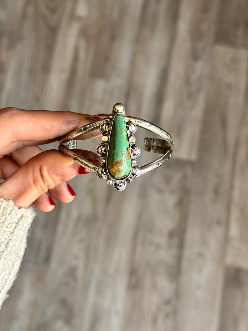 May include: Silver cuff bracelet featuring a large, teardrop-shaped turquoise stone. The stone is set in a decorative bezel with ornate silver detailing. The bracelet has a hinged design for easy wear and is a statement piece of jewelry.
