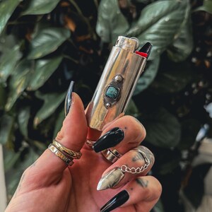 May include: A silver lighter with a turquoise stone inlay. The lighter is being held in a hand with black nail polish.