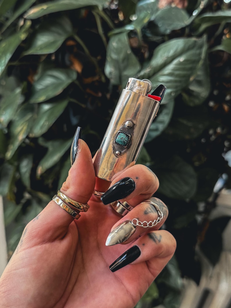 May include: A silver lighter with a turquoise stone inlay. The lighter is being held in a hand with black nail polish.