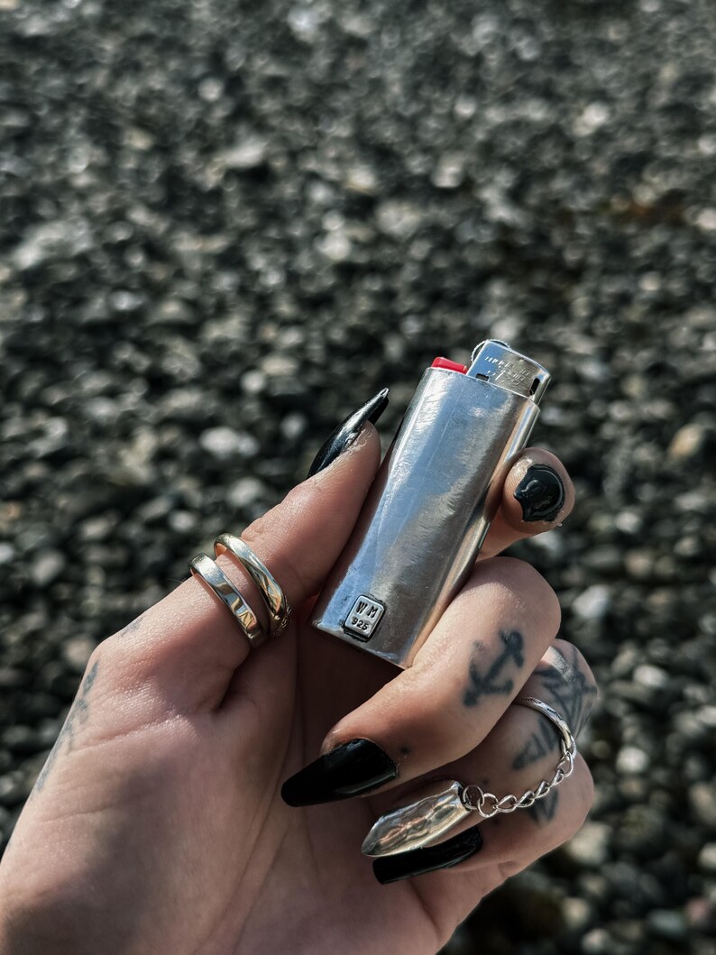 May include: A silver lighter with a red top and the text "WM 925" on the side. The lighter is being held by a hand with black nail polish.